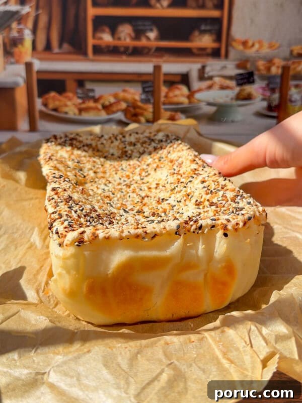 Viral Blender Rice Bread Recipe — Gluten-Free, No Flour 4 A hand holds a rectangular, seeded bread—reminiscent of the viral Blender Rice Bread—covered in sesame and poppy seeds, resting on parchment paper with a bakery display of breads and pastries in the background.