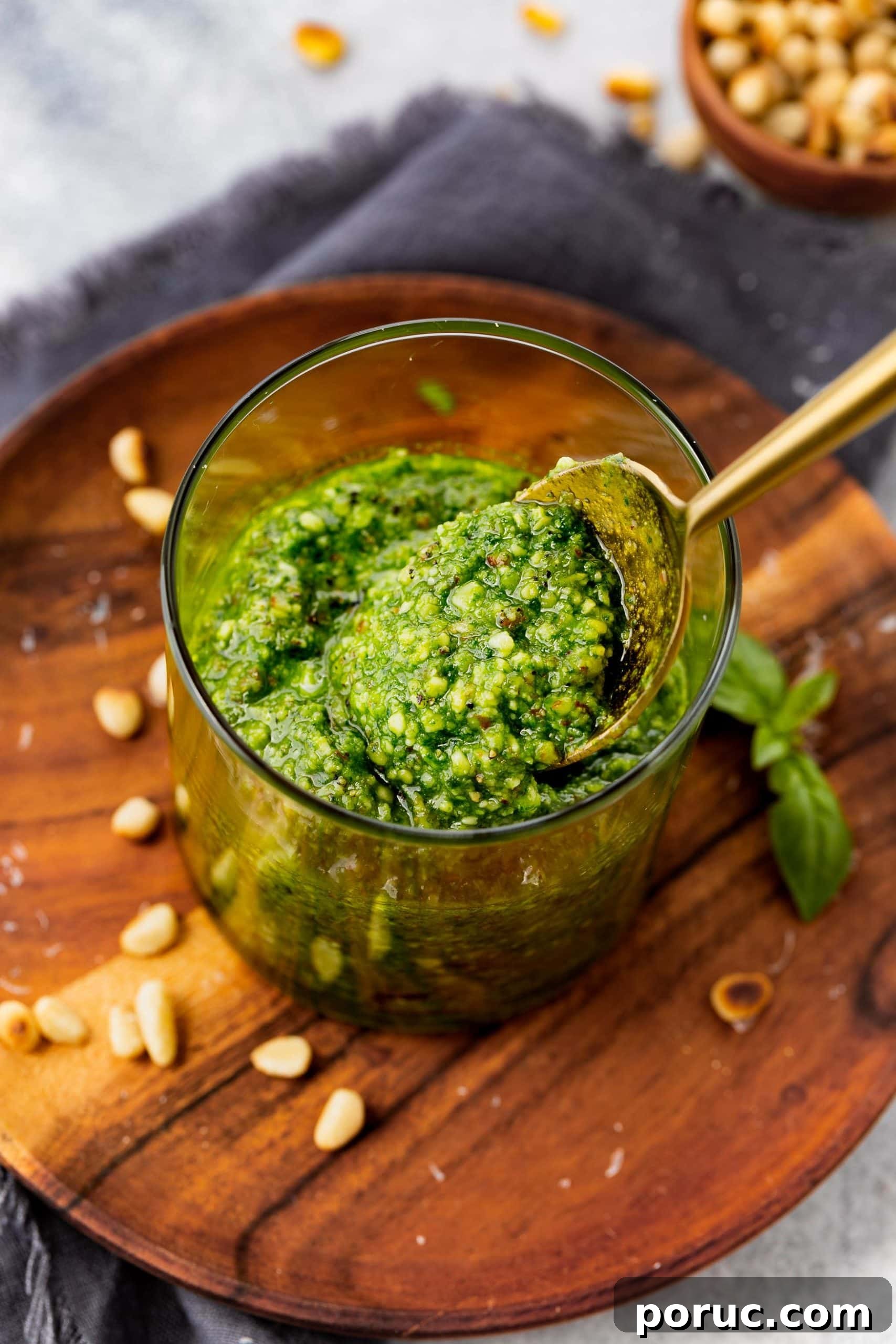 Homemade Classic Basil Pesto Recipe for Pasta & Sandwiches 4 a photo of a jar of pesto sauce with a gold spoon scooping some out. the jar is on a wooden plate and has some pine nuts and fresh basil leaves on it.
