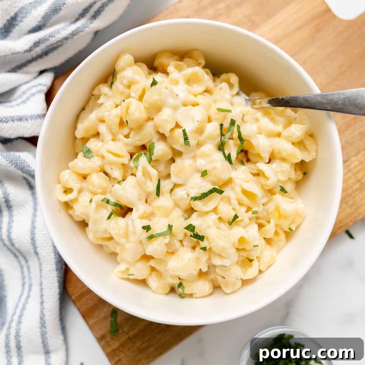 A bowl of creamy macaroni and cheese garnished with chopped herbs, with a fork resting in the bowl on a wooden board.