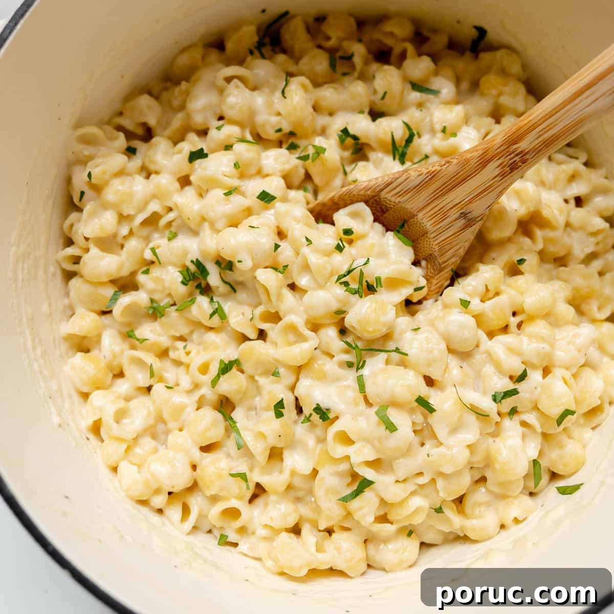 A pot of creamy macaroni and cheese garnished with chopped herbs, with a wooden spoon resting in the mixture.