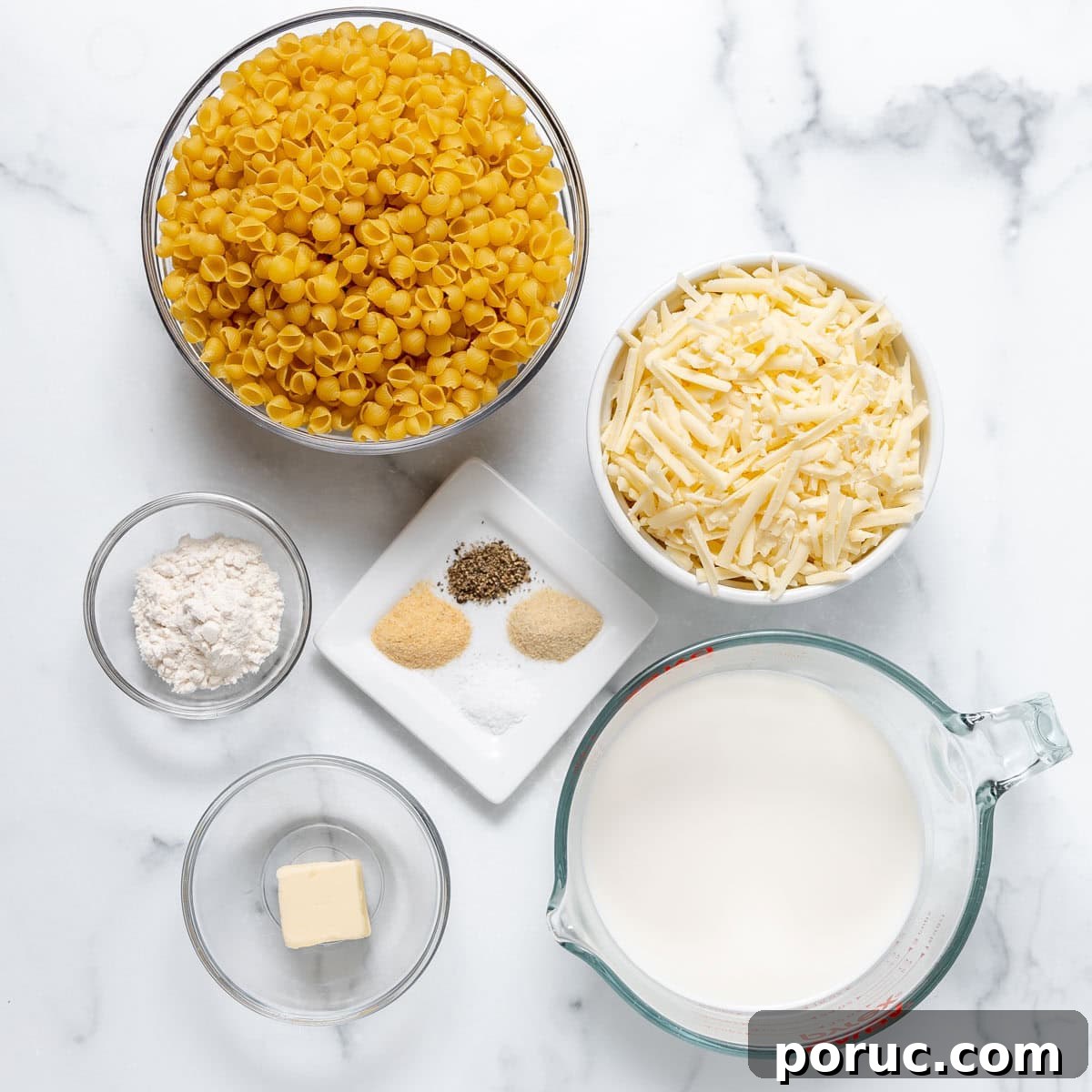 Top-down view of bowls containing dry pasta, shredded cheese, flour, butter, milk, and spices on a white marble surface.