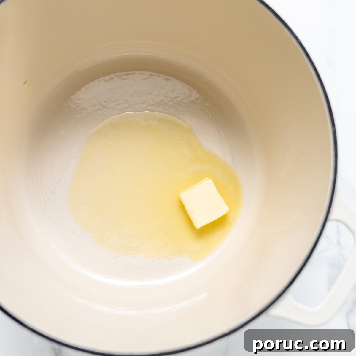 A small cube of butter partially melted in a large cream-colored pot on a white marble surface.