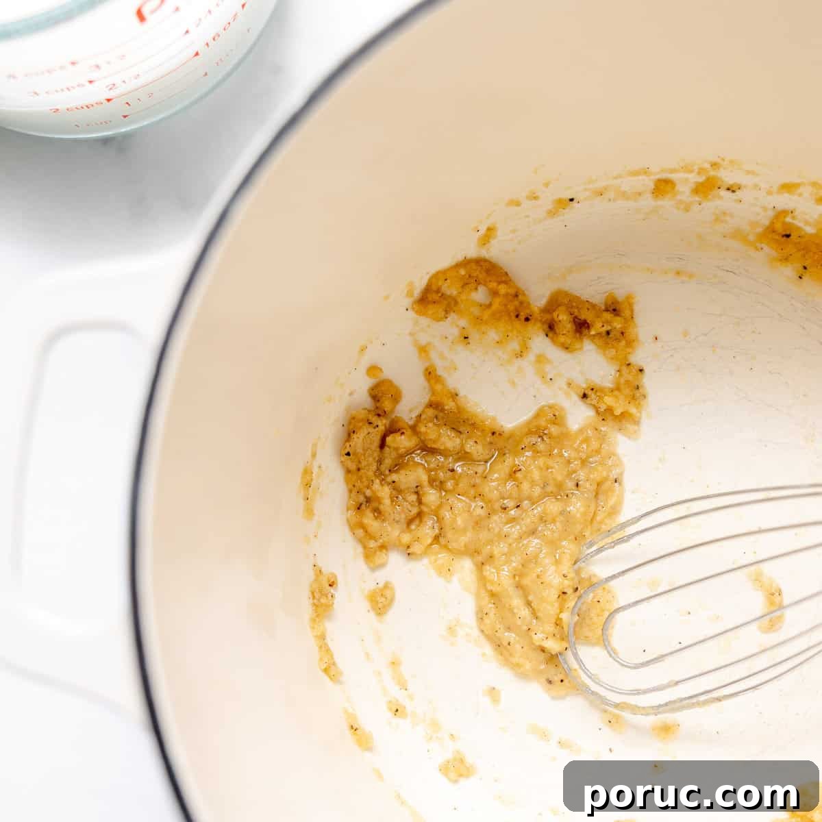 A whisk rests in a white pot containing a small amount of cooked roux. A glass measuring cup is partially visible in the corner.