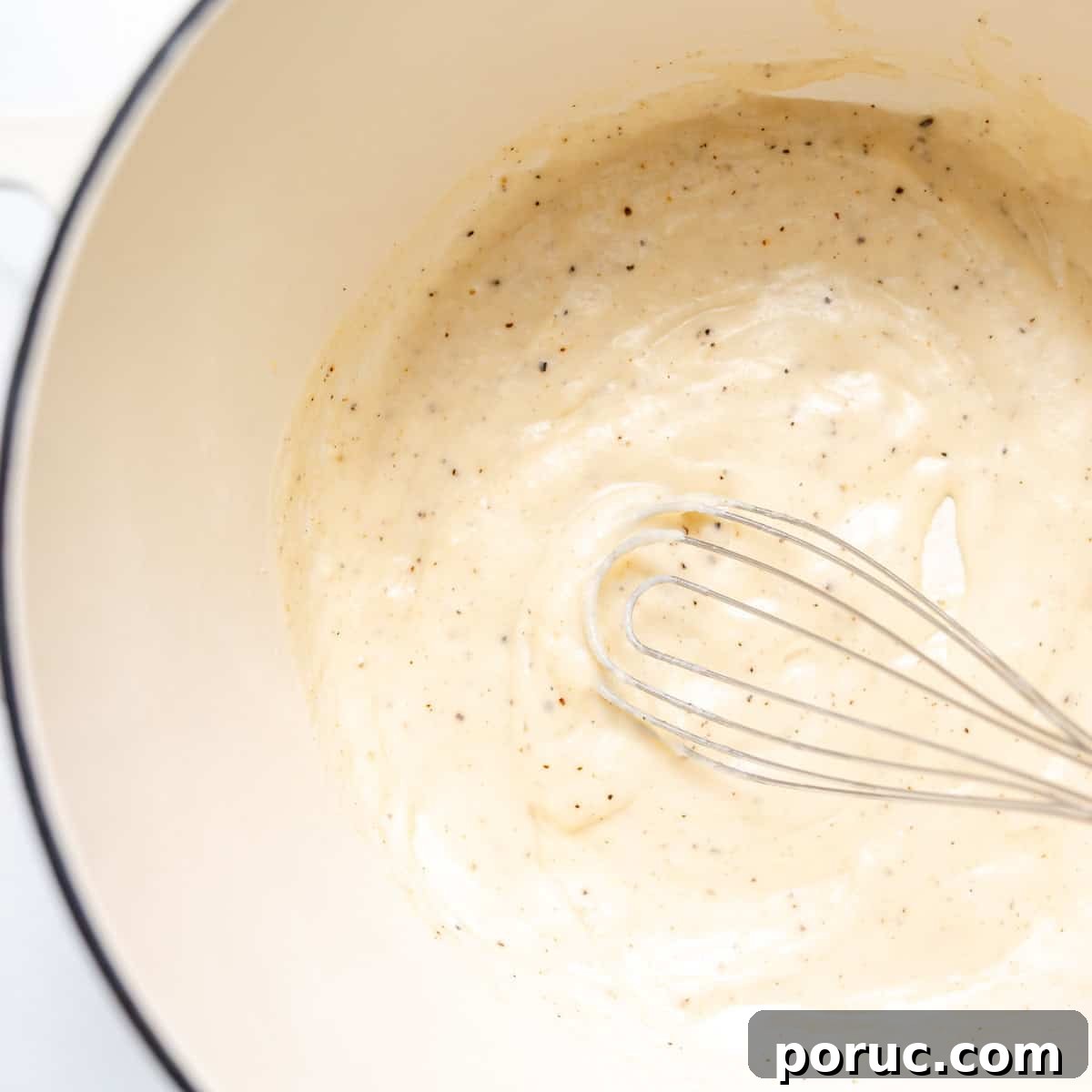 A metal whisk stirring a creamy white sauce with black pepper flecks in a white mixing bowl.