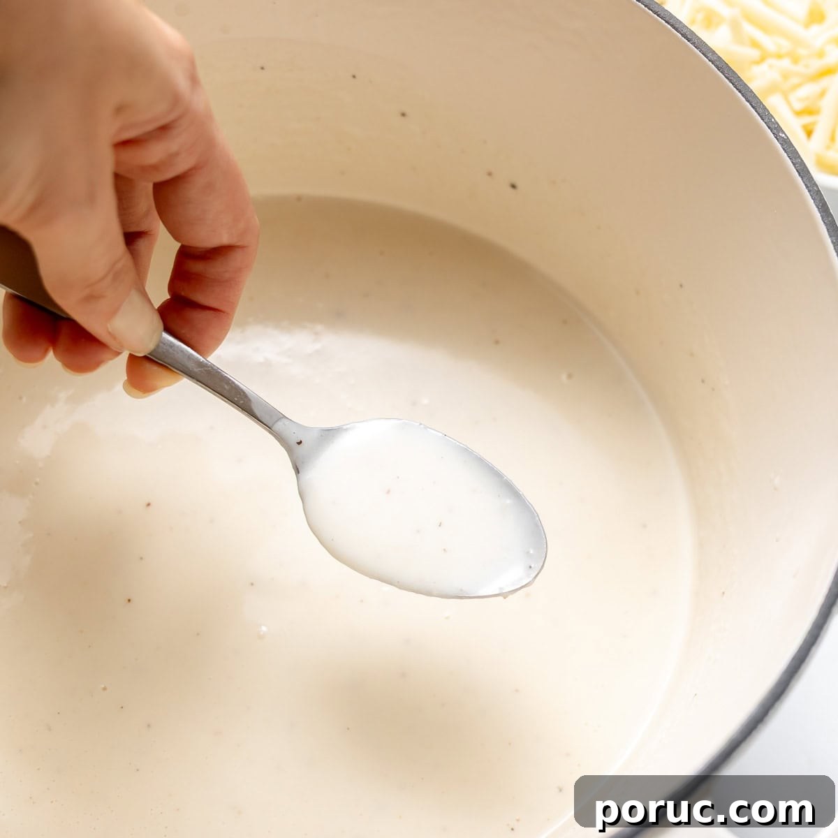 A hand holds a spoon with a white creamy sauce over a pot containing more of the same sauce.