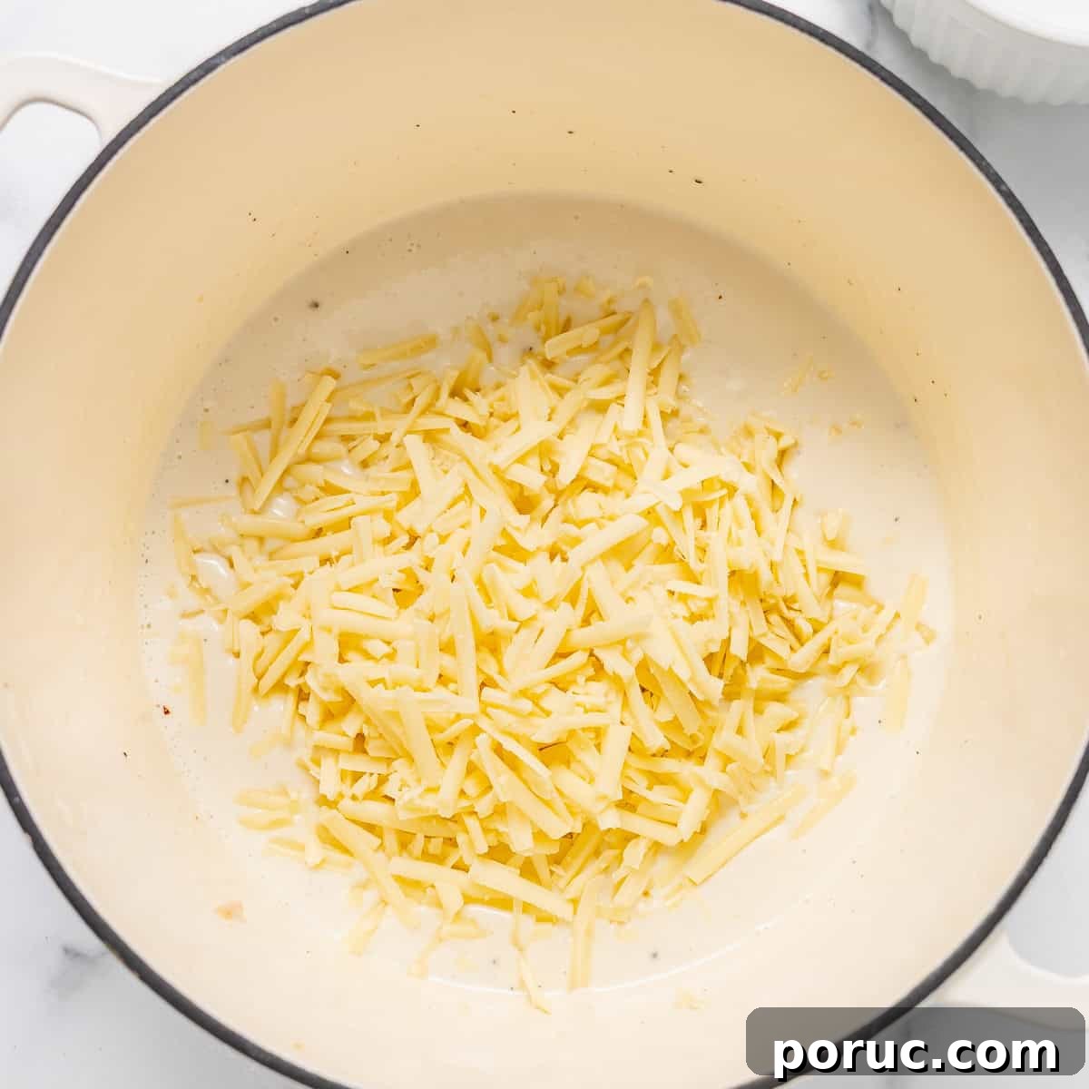 Shredded cheese in a cream-colored pot, ready to be melted, with a small white ramekin visible in the background.