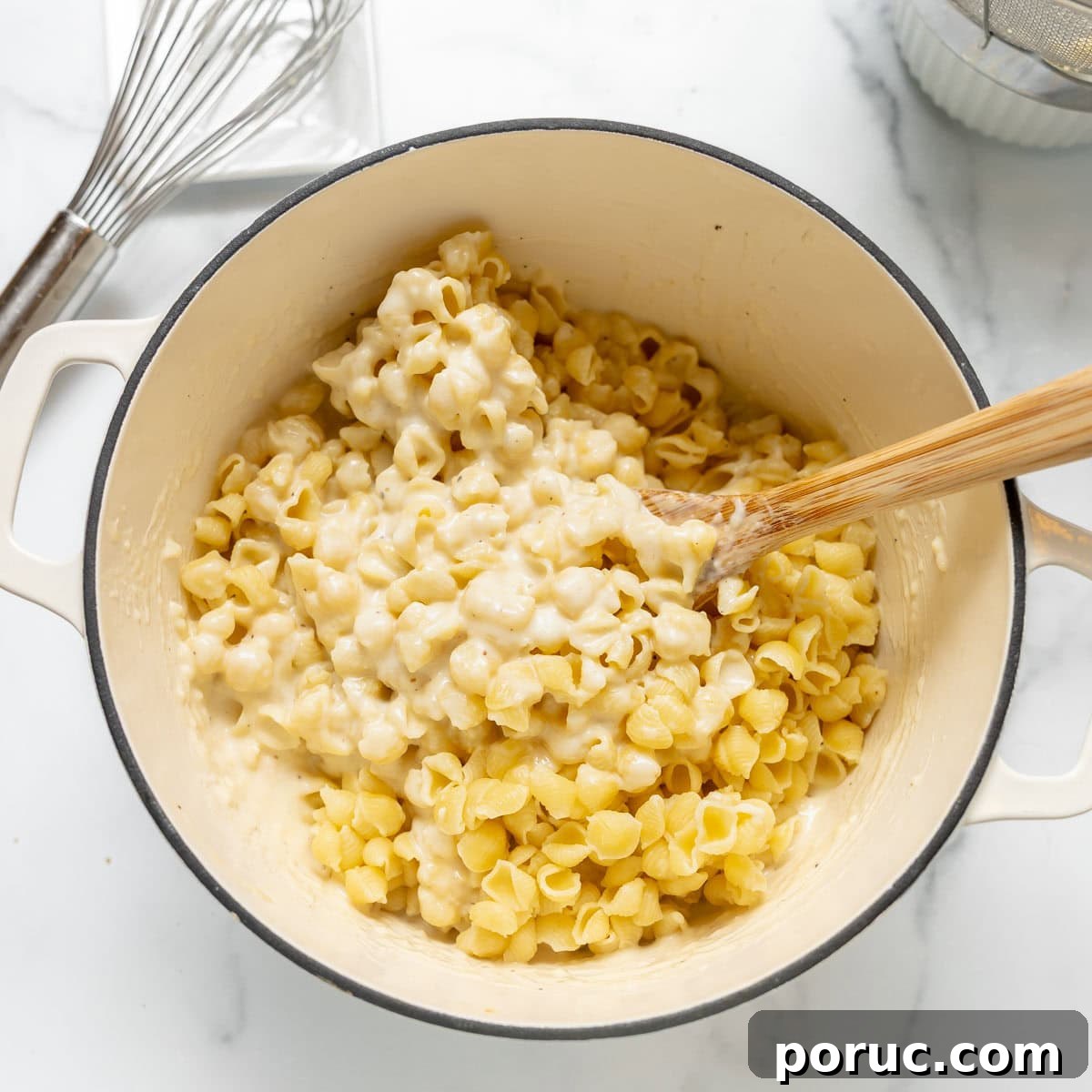 A pot of cooked pasta shells mixed with creamy cheese sauce, being stirred with a wooden spoon on a white countertop.