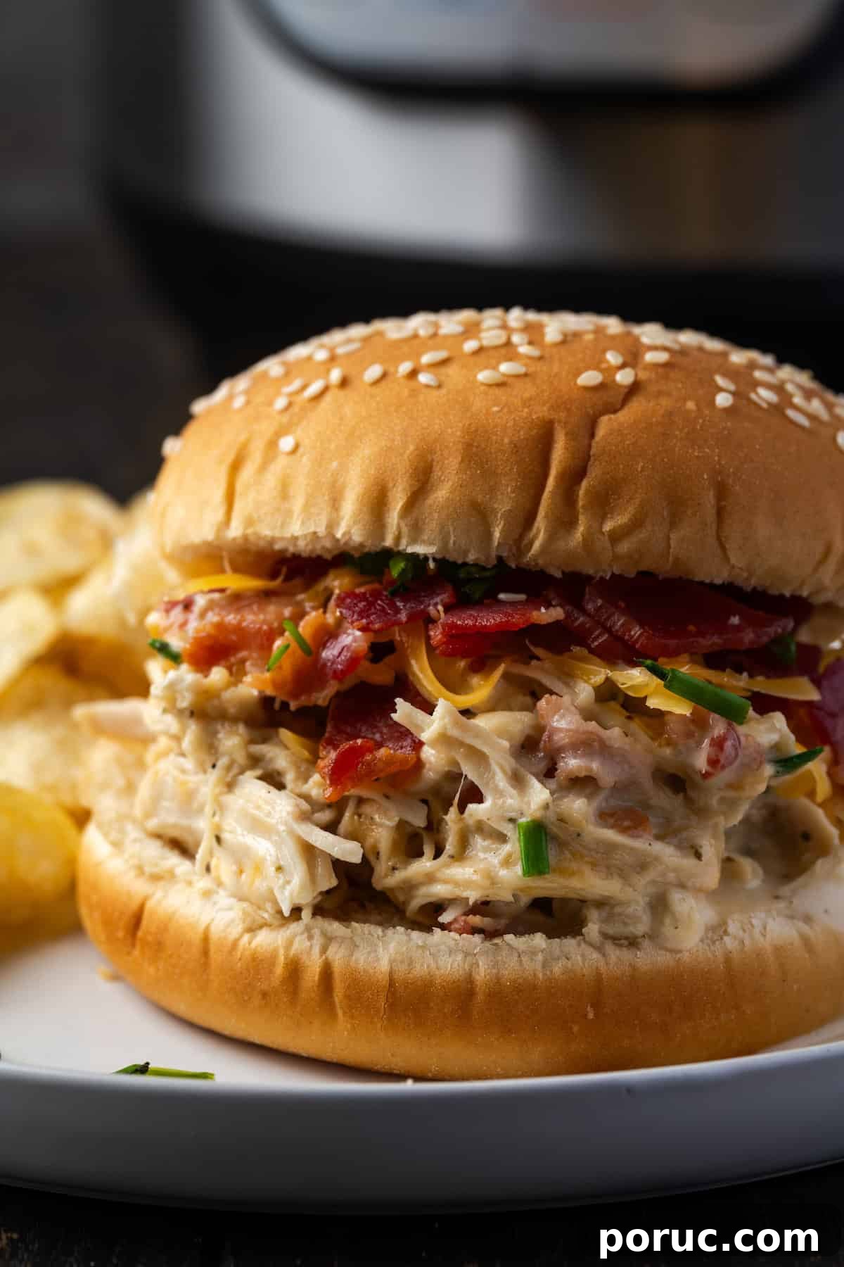 Close up of a crack chicken sandwich on a plate next to potato chips, with the Instant Pot in the background.